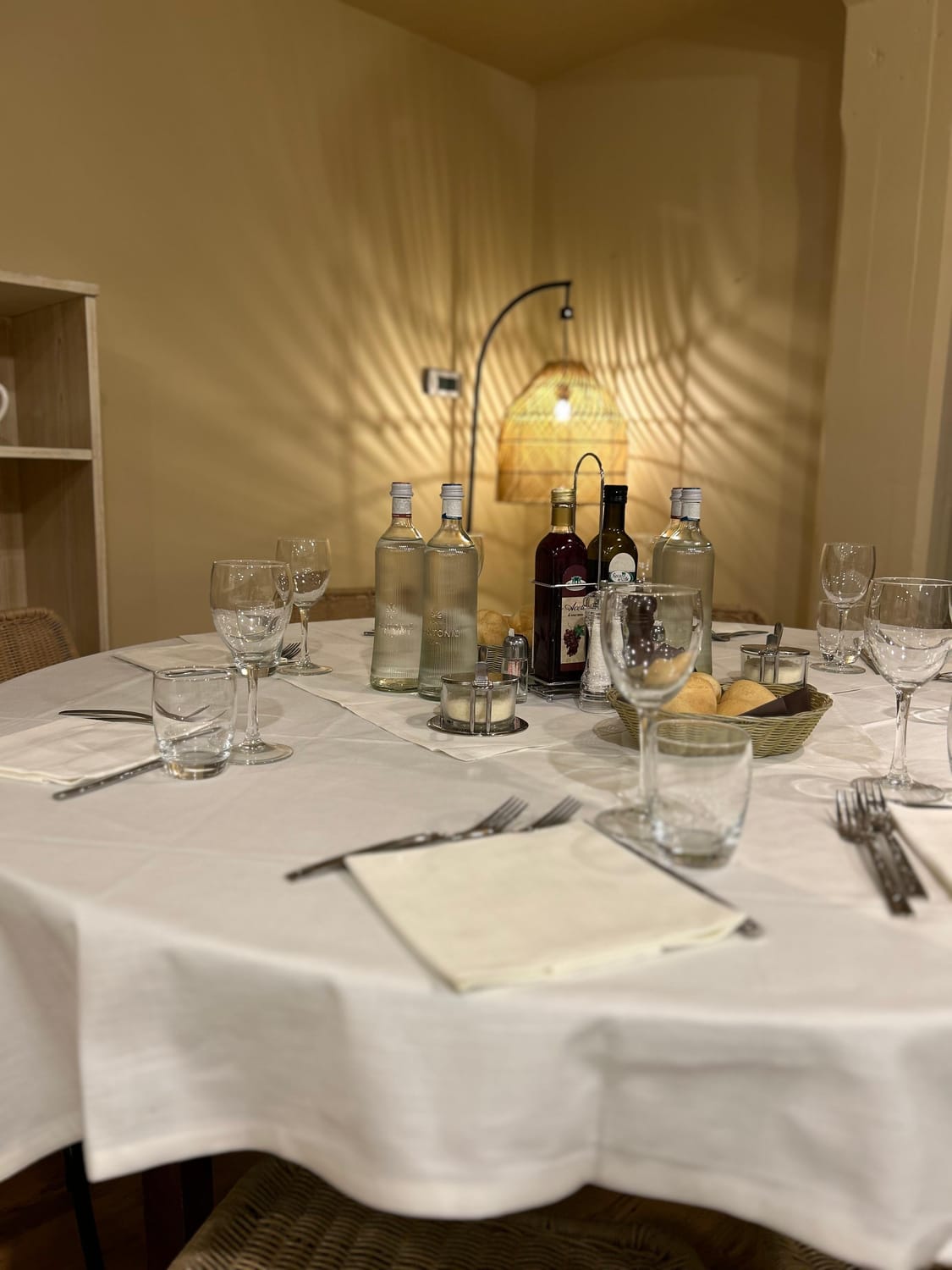 Tavola apparecchiata con bicchieri, acqua, olio, aceto e un cestino di pane, illuminata da una lampada in vimini
